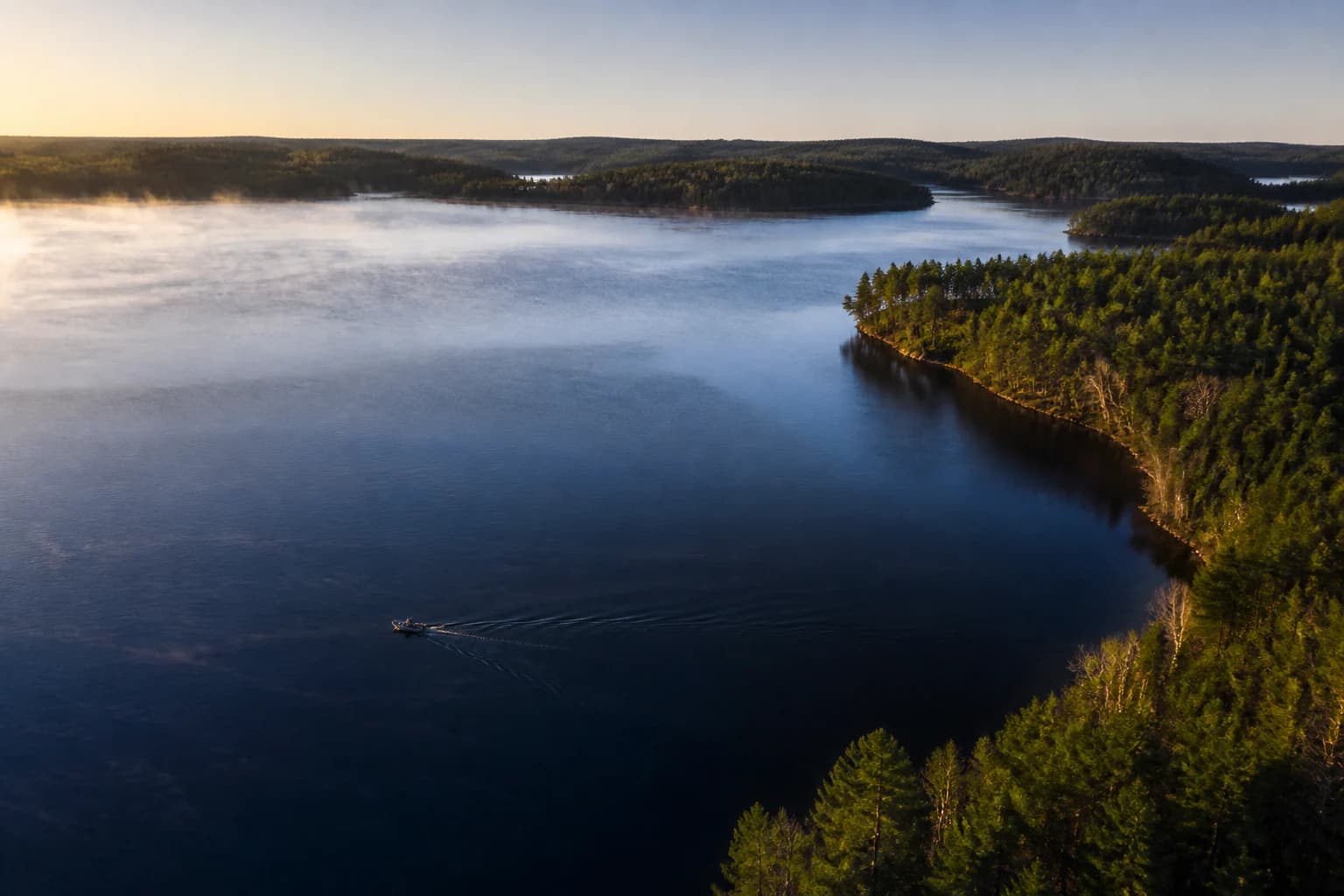 A small fishing boat crossing a broad forest-lined lake at first light